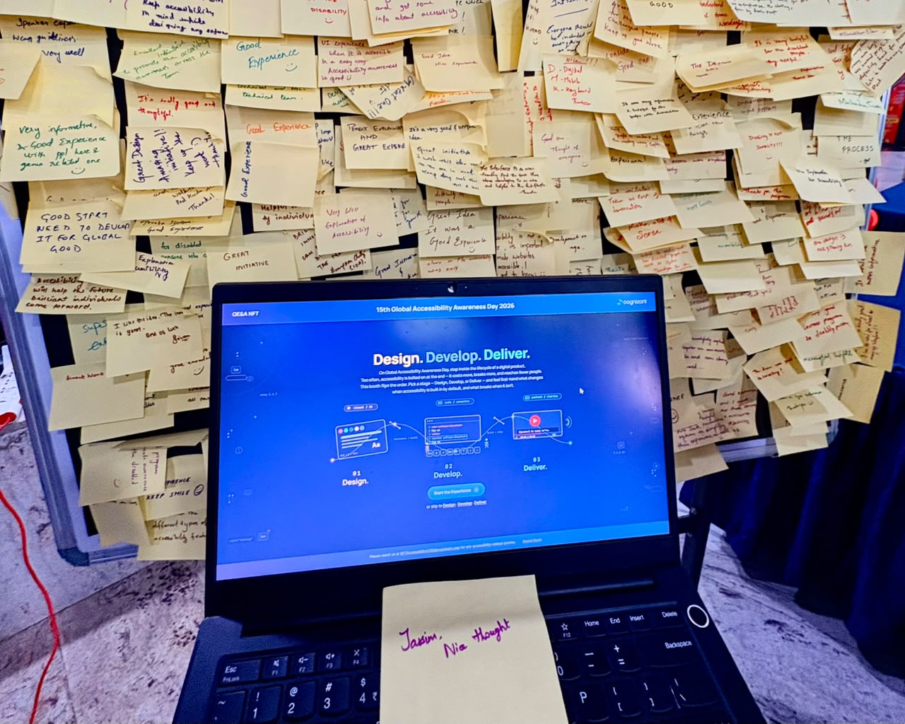 The GAAD 2026 booth in action: an open laptop running the kiosk's blue 'Design. Develop. Deliver.' start screen, surrounded by a wall of handwritten yellow feedback sticky notes left by visitors.
