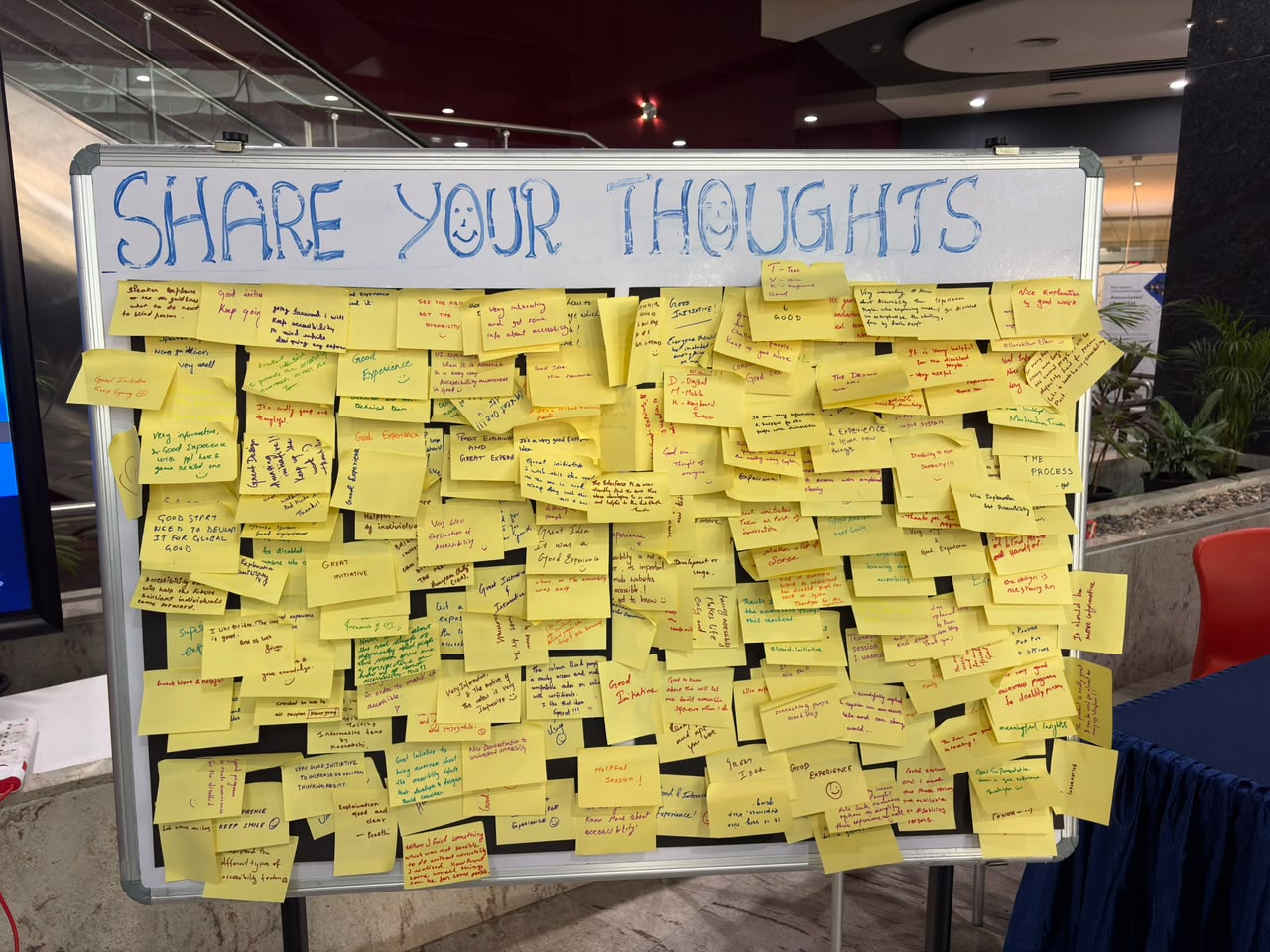 A whiteboard headed 'Share Your Thoughts' (with a smiley in the O) at the GAAD 2026 booth, covered with dozens of handwritten yellow sticky notes of visitor feedback in the event hall.